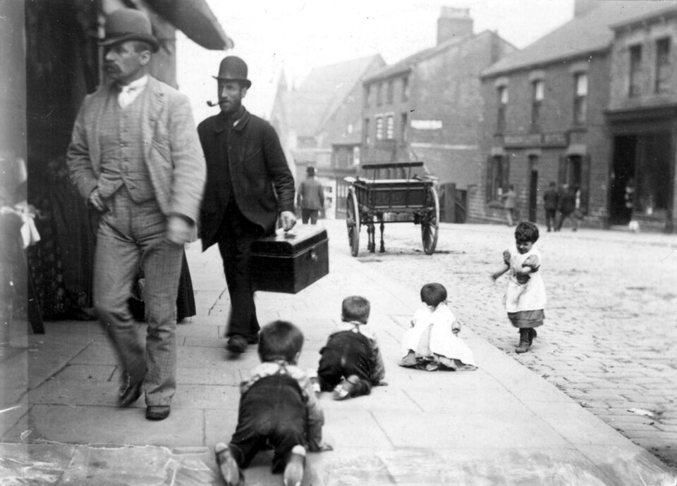 A black and white photograph of a street scene from the past. In the foreground, four children are sitting and playing on the pavement.